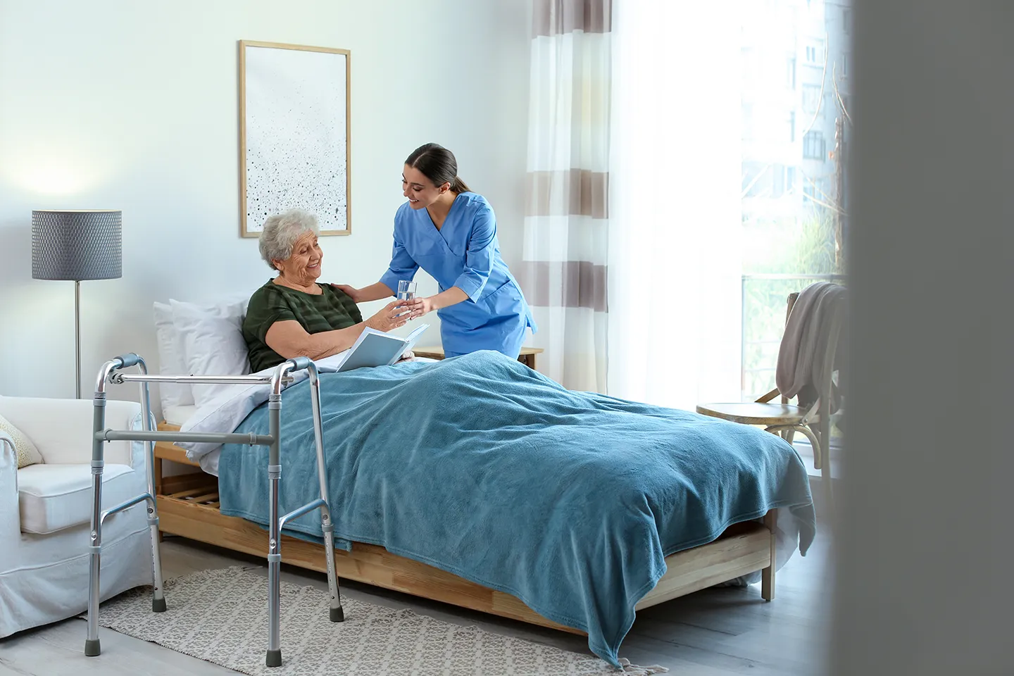 A nurse in blue scrubs assists an elderly woman sitting in a hospital bed, with a walker nearby in a bright, well-lit room.