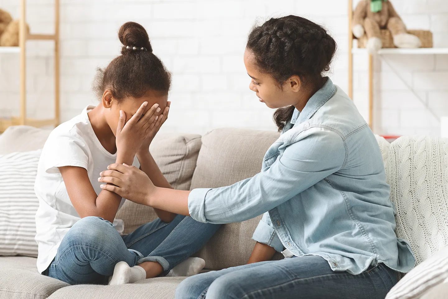 A girl comforts another girl who is sitting on a couch, covering her face with her hands. They both wear casual clothes in a cozy room.