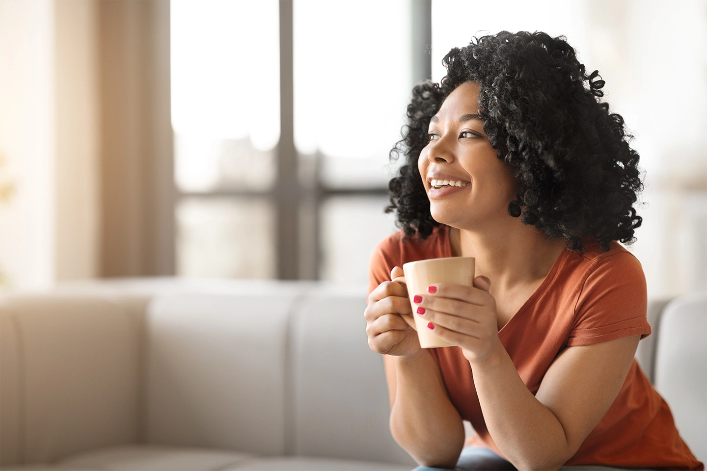 Woman with curly hair smiling and holding a mug, sitting on a sofa in a sunlit room.