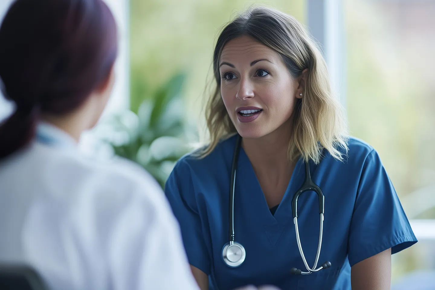 Two healthcare professionals in conversation, one wearing blue scrubs with a stethoscope, the other in a white coat, in a bright setting.